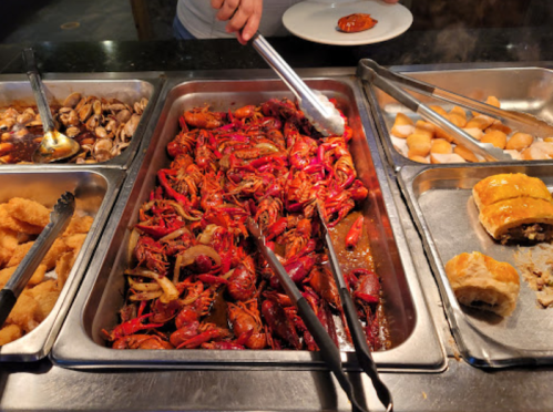 A buffet tray filled with spicy crawfish, surrounded by various seafood dishes and tongs for serving.