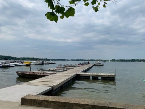 A wooden dock extends into a calm lake, lined with boats under a cloudy sky. Green leaves frame the scene.