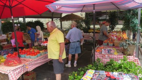 A bustling farmers market with colorful produce, shoppers browsing under red umbrellas, and tables covered in checkered cloth.