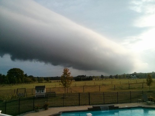 A dramatic cloud formation looms over a grassy field, with a pool and fence in the foreground.