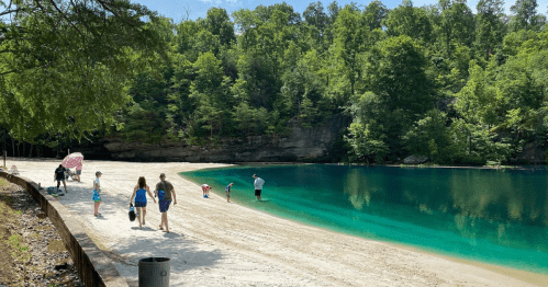 A serene lake surrounded by lush greenery, with people walking along the sandy shore and wading in the water.