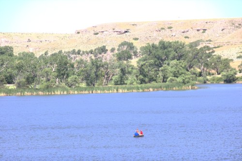 A small boat with two people floats on a calm lake surrounded by green trees and rolling hills.