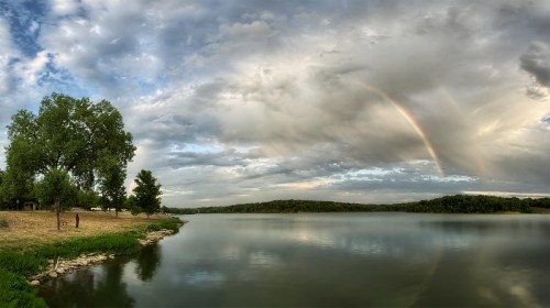 A serene lake scene with trees, a rainbow in the sky, and clouds reflecting on the water's surface.