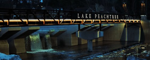 A bridge over a waterway at night, illuminated with the sign "Lake Peachtree" and flowing water beneath.