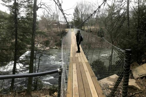A person stands on a wooden suspension bridge over a river, surrounded by trees and a serene landscape.