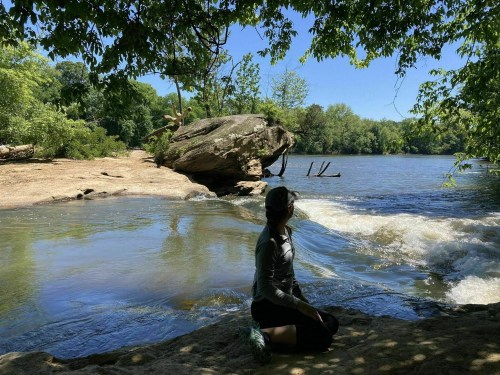 A person sits by a river, surrounded by trees, with a rocky shore and gentle waves under a clear blue sky.