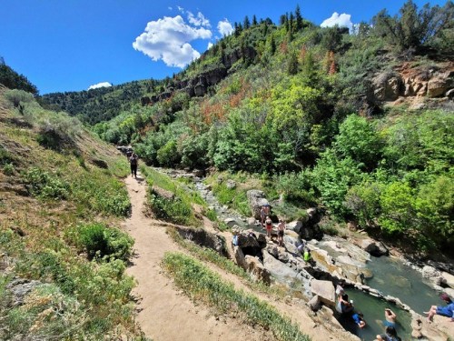 A scenic trail alongside a stream, surrounded by lush greenery and rocky terrain under a bright blue sky.