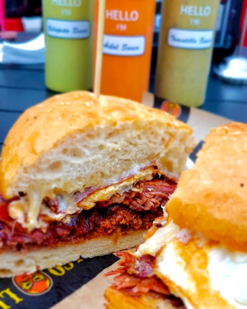 Close-up of a delicious sandwich with layers of meat and cheese, served with colorful sauces in the background.