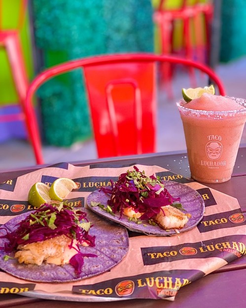 Two tacos with fish, purple cabbage, and lime wedges on a table, alongside a margarita in a glass with salt on the rim.