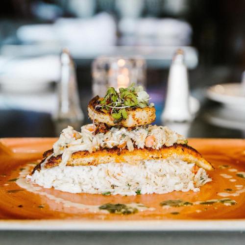 A beautifully plated dish featuring grilled fish atop crab meat and rice, garnished with microgreens on an orange plate.