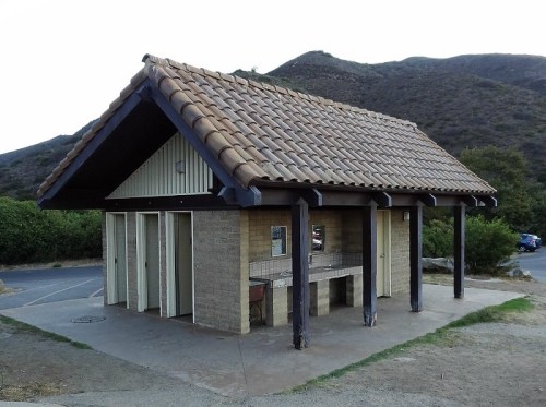 A small, rustic restroom building with a tiled roof, set against a backdrop of hills and greenery.