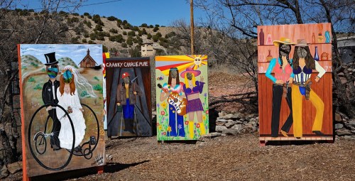 Four colorful paintings displayed outdoors, featuring whimsical characters in various outfits and settings.