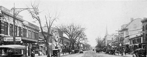 Historic street scene with vintage cars, storefronts, and bare trees in a snowy setting.