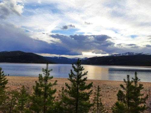 A serene lake surrounded by mountains, with a cloudy sky and pine trees in the foreground.
