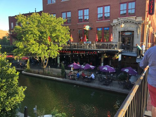 A riverside restaurant with outdoor seating, surrounded by greenery and red flags, under a clear blue sky.
