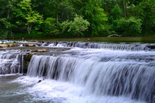 A serene waterfall cascades over rocky ledges, surrounded by lush green trees and a calm river.