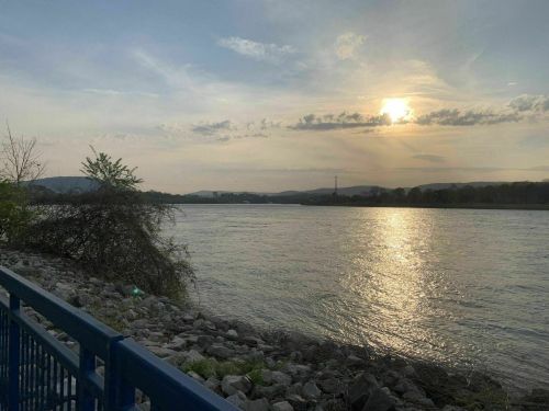 Sunset over a river, with calm waters reflecting the sky and distant hills, framed by trees and a blue railing.