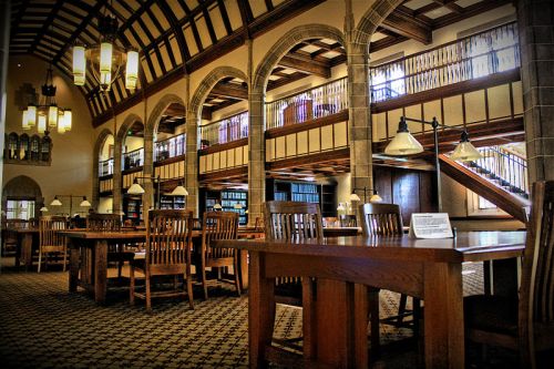 A spacious library interior featuring wooden tables, arched windows, and a high ceiling with decorative lighting.
