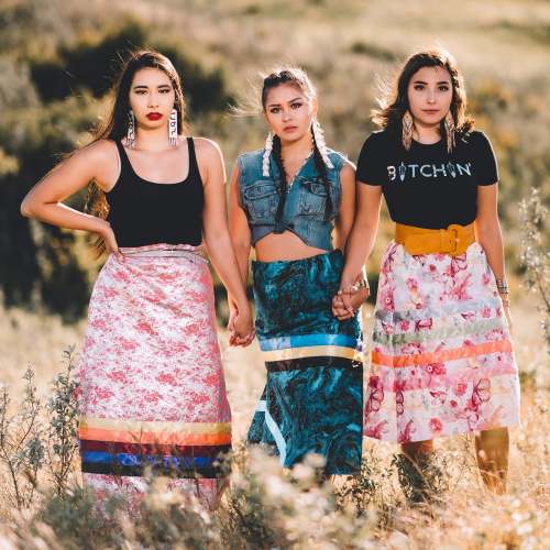 Three women stand together in a field, wearing colorful skirts and stylish tops, showcasing a vibrant, confident look.