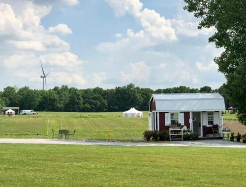 A small red shed with a white roof sits in a green field, with a wind turbine and trees in the background under a cloudy sky.