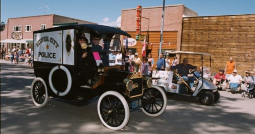 A vintage police car and a golf cart parade through a town, with spectators watching from the sidelines.