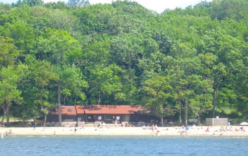A sandy beach with people enjoying the sun, surrounded by lush green trees and a rustic building in the background.