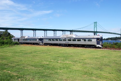 A vintage train travels along tracks near a green field, with a large bridge and blue sky in the background.