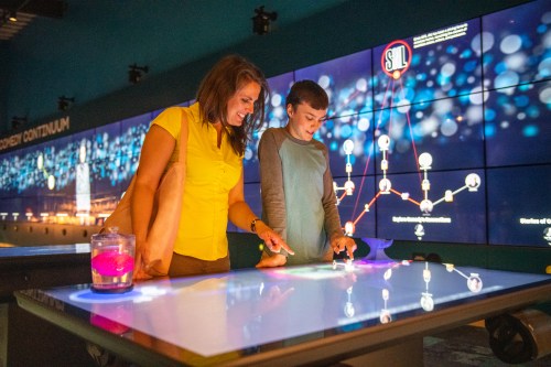 A woman and a boy interact with a touchscreen display in a modern exhibit, colorful lights in the background.