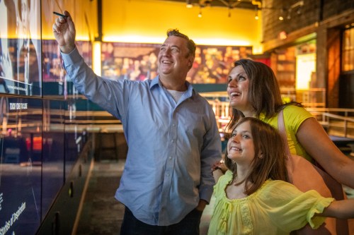 A man, woman, and girl smile and point at an interactive display in a museum.