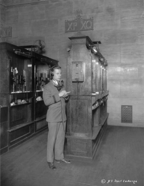 A man in a suit stands by vintage telephone booths in a historic setting, wearing a headset and holding a device.