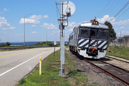 A train on tracks beside a road, with a signal pole nearby and a scenic view of water in the background.