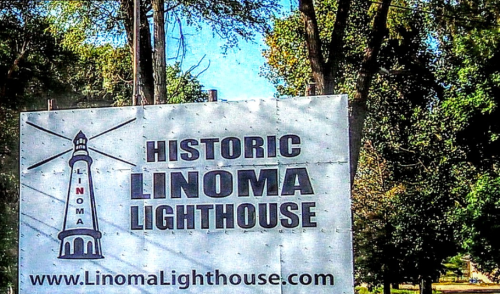 Sign for Historic Linoma Lighthouse, featuring a lighthouse graphic and website link. Surrounded by trees.