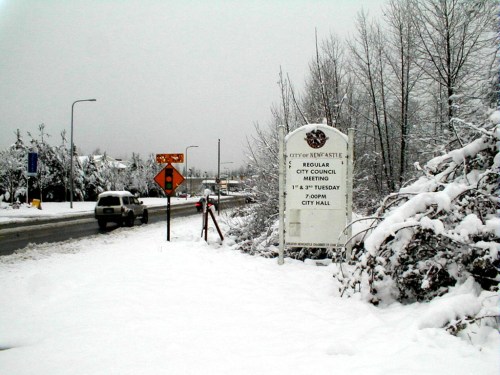 A snowy street scene featuring a sign for a city council meeting at City Hall in New Westminster.