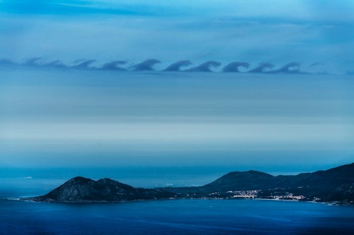 A serene coastal landscape featuring a calm sea, distant hills, and unusual cloud formations resembling waves in the sky.