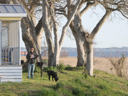 A person stands near a house, holding a drink, with a black dog beside them and trees in the background by a lake.