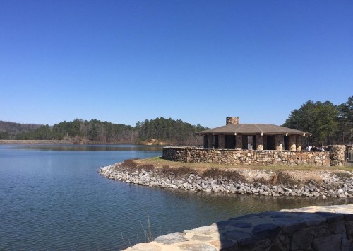 A stone pavilion by a calm lake, surrounded by trees and clear blue skies.