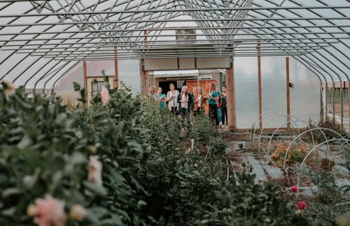 A group of women stands inside a greenhouse, surrounded by lush plants and flowers, smiling and engaging with each other.