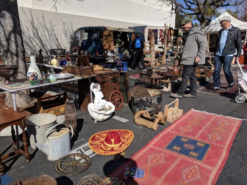 A bustling outdoor market with various antiques and vintage items displayed on tables and the ground. Two people walk by.