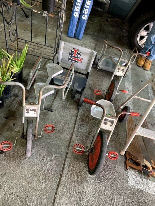 Three vintage tricycles with red pedals and wheels, displayed on a concrete surface alongside plants and a vehicle.