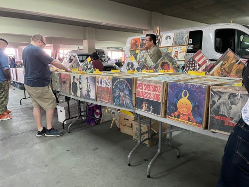 A bustling market scene with people browsing a table filled with vinyl records and music memorabilia.