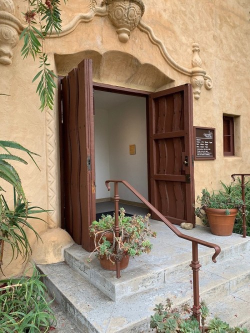 A wooden door opens to a light interior, surrounded by plants and decorative stonework. Steps lead up to the entrance.