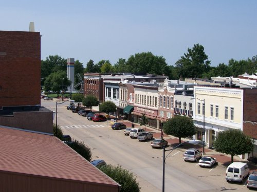 A view of a small town street lined with historic buildings, trees, and parked cars under a clear blue sky.