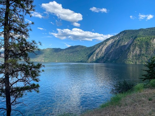A serene lake surrounded by mountains and trees under a blue sky with fluffy clouds.