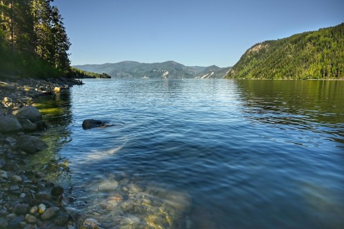 A serene lake surrounded by mountains and trees, with clear water reflecting the blue sky.