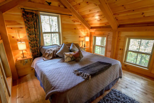 Cozy wooden bedroom with a large bed, decorative pillows, and windows overlooking a forested area.
