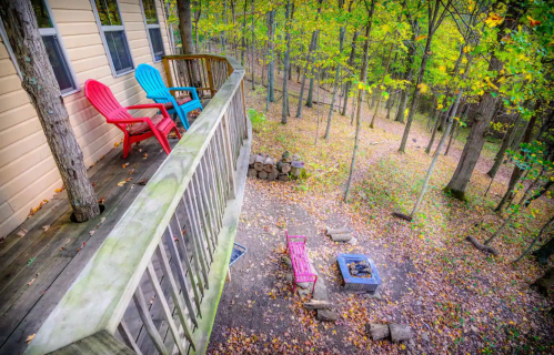 A wooden deck with colorful chairs overlooks a forested area with a fire pit and fallen leaves.