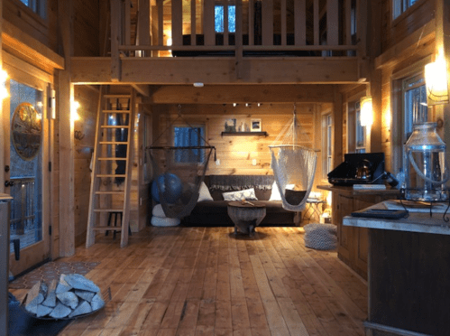 Cozy wooden cabin interior with a sofa, hammocks, and warm lighting, featuring a ladder and a rustic fireplace.