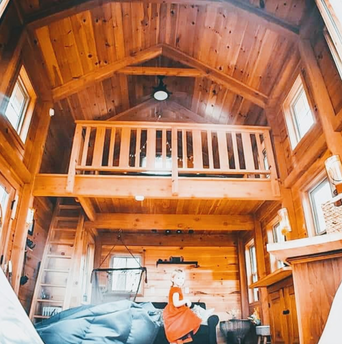 Cozy wooden interior of a tiny house with a loft, featuring a child in a red dress sitting on a couch.