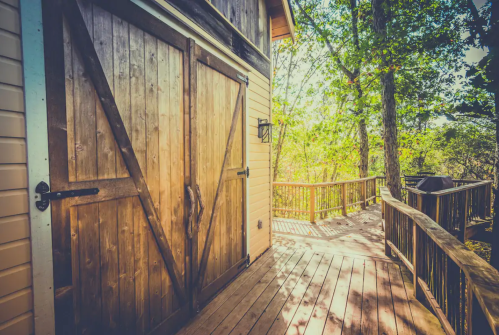 A wooden deck leads to a rustic barn-style door, surrounded by trees and natural light.
