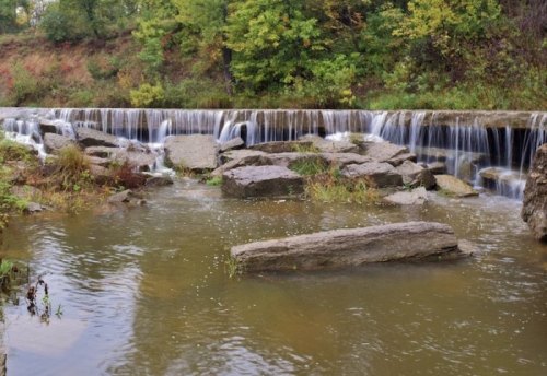 A serene waterfall cascades over rocks into a calm river, surrounded by lush greenery and autumn foliage.